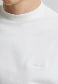 Weißes Baumwoll-T-Shirt mit hohem Hals und dezentem "Prohibited"-Stickereien in passender Farbe, das eine glatte Textur präsentiert.
