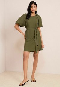 Rochie verde oliv cu guler rotund, mâneci bufante și curea asortată. Textură netedă, croială până la genunchi, asociată cu sandale negre.
