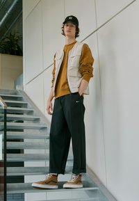 Tenisi bej cu talpă albă, pantaloni negri cu logo-ul Nike, hanorac bej și o vestă crem cu multiple buzunare, plasate într-un mediu cu o scară modernă.