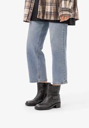 Personne portant un jean bleu court, des bottines en cuir noir avec boucle, une veste à carreaux et une chemise noire, debout sur un fond blanc.