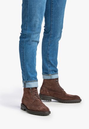 Bottines en daim marron avec des détails en brogue, associées à un jean slim bleu avec des poignets retroussés. Semelle en caoutchouc noir pour une meilleure adhérence.