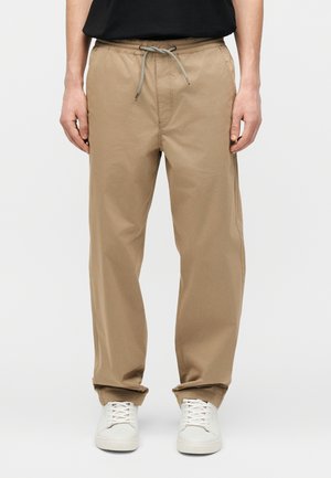 Bărbat purtând pantaloni bej cu șnur și adidași albi, stând în fața unui fundal simplu.