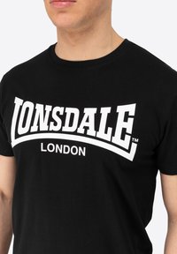 Juodas medvilninis marškinėliai su ryškiu baltu "LONSDALE" logotipu ir "LONDON" tekstu, parašytu drąsiu, išlenktu šriftu per krūtinę.