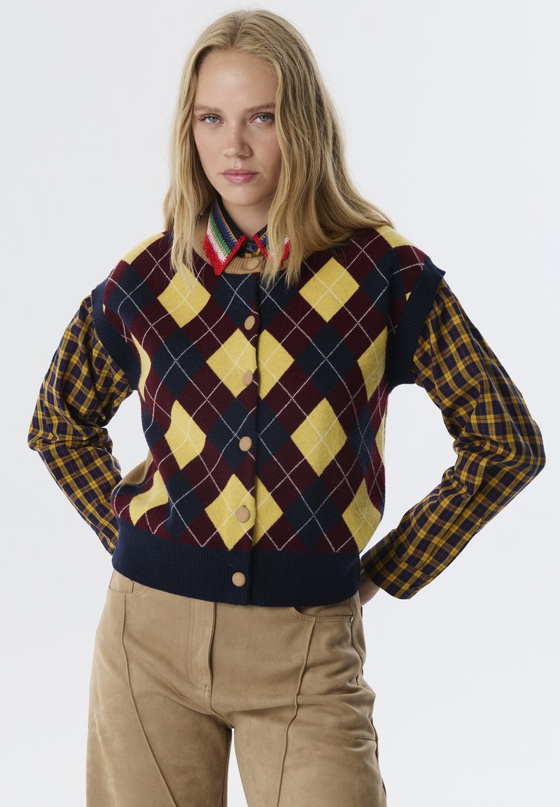 Strickjacke mit navyblauem, gelbem und burgunderfarbenem Argyle-Muster; runde Knöpfe und gerippter Saum; kombiniert mit einem karierten Hemd.