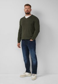 Dunkelgrüner V-Ausschnitt-Pullover, blaue Jeans und beigetöne Sneakers mit weißen Akzenten. Schlichtes Design mit strukturierter Oberfläche.