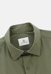 Chemise à col olive avec fermeture à boutons, texture lisse, et une étiquette portant l'inscription "Serge Blanco" centrée sous le col.