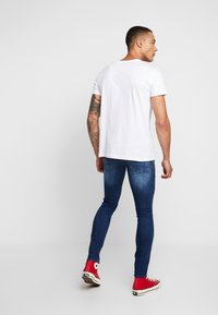 Man med tatuerad arm, klädd i vit t-shirt, blå skinny jeans och röda höga sneakers, stående mot en enkel vit bakgrund.
