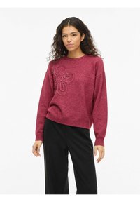 Pull couleur marron avec un motif floral texturé sur le devant, doté d'un col rond et de poignets côtelés, associé à un pantalon noir.