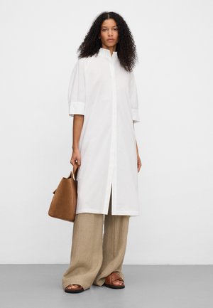 Donna che indossa un vestito camicia bianco lungo con bottoni, pantaloni beige a gamba larga, sandali marroni a ciabatta e tiene in mano una borsa marrone.