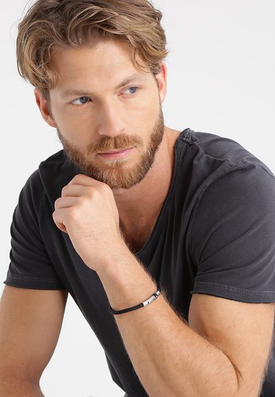 Black t-shirt, close-fitting. Model wearing a braided black bracelet with silver accents. Light skin, short wavy hair, beard.