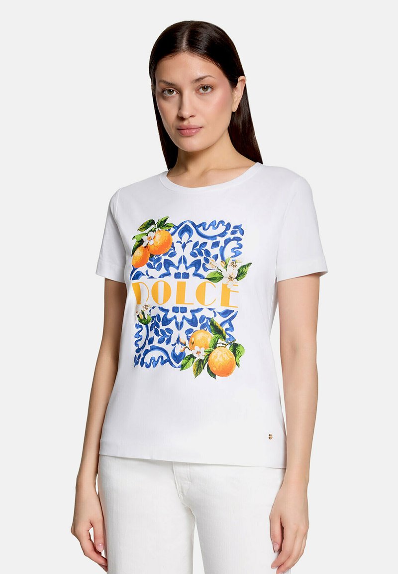 Femme portant un t-shirt blanc à manches courtes avec un motif floral bleu, des illustrations de fruits oranges et le mot « DOLCE » en lettres jaunes.