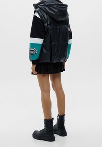 Veste noire rembourrée avec capuche, dotée de panneaux multicolores, d'un écusson logo sur la manche et d'une texture lisse. Portée sur une jupe plissée noire et des bottes noires.