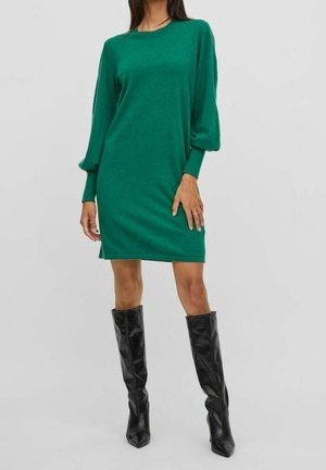 Kvinde iført en grøn knælængde kjole med lange ærmer og manchetter samt sorte knæhøje støvler med spidst tåparti.