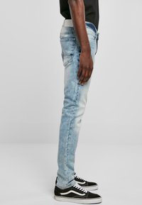 Los jeans de mezclilla azul claro presentan un corte ajustado, un acabado desgastado y cinco bolsillos. Una camisa negra es parcialmente visible, junto con unas zapatillas negras y blancas.