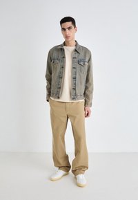 Jeansjacke in hellblau mit Knopfverschluss, darunter ein beige Rundhals-T-Shirt und lockere beige Hose. Weiße Sneakers runden das Outfit ab.