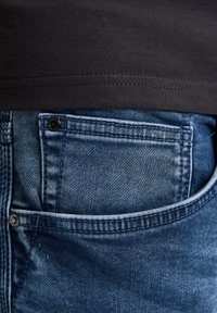 Modré džíny z denimu s kapsou na zadní straně, s tmavým šitím, nýtem a texturovaným povrchem. Černá látka viditelná nad pasem.