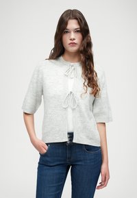 Gai pelēks kardigans ar īsām piedurknēm, diviem priekšējiem saišu galiem un ribotu apakšmalu. Valkāts virs baltas blūzes ar tumši zilām džinsām. Mīksta tekstūra.