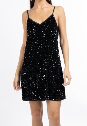 Kobieta w czarnej, mini sukience bez rękawów z cienkimi ramiączkami, pokrytej błyszczącymi cekinami, widoczna od ramion do połowy uda.