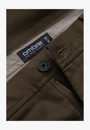 Pantaloni in cotone marrone con motivo a spina di pesce, dotati di un bottone nero, cerniera argentata e un'etichetta con la scritta "ombre quintessence M:32."