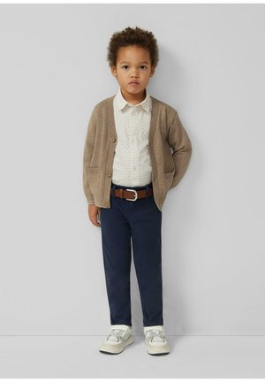 Małe dziecko o kręconych włosach, ubrane w beżowy kardigan, wzorzystą koszulę, granatowe spodnie, brązowy pasek i białe trampki, stoi na tle szarości.