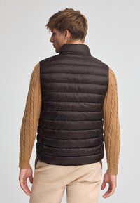 Wypchana ciemno-brązowa kamizelka z wysokim kołnierzem, w wzór pikowanej tkaniny. Noszona na beżowym swetrze z warkoczem, zestawiona z jasnymi beżowymi spodniami.