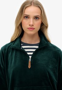 Sudadera de forro polar verde oscuro con cuello con cremallera, que presenta una capa interior estampada, logo bordado y acento de etiqueta de cuero.