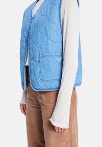 Blauw gewatteerd vest met een zak, voorzien van een subtiel hartdetail. Draagt over een witte, met patroon bedrukte, lange mouw shirt en gecombineerd met bruine ribfluwelen broek.