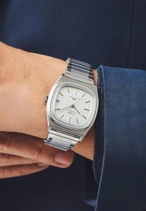 Srebrna kovinska ura s kvadratnim zaščitnim steklom, belo številčnico, srebrnimi oznakami in nerjavečim jeklenim paščkom, nosena na zapestju z modrim rokavom srajce.