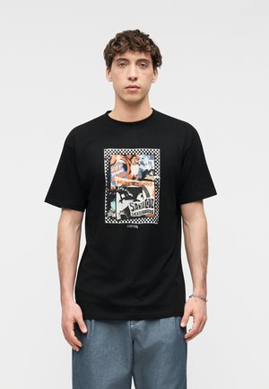 Tânăr purtând un tricou negru cu un grafic colorat de skateboard și textul "Santa Cruz Skateboards", asortat cu blugi albaștri.