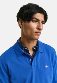 Blauwe polo van een gestructureerde stof, met een klassieke kraag en logodetail. Draagt over een geruite overhemden met knopen.