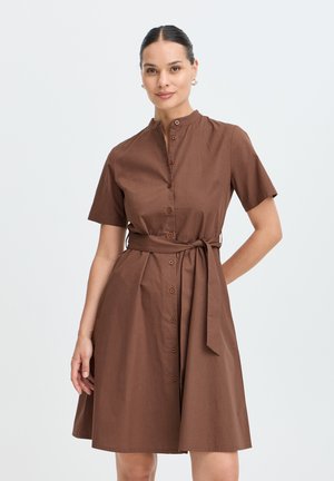 Femme aux cheveux foncés portant une robe marron à manches courtes boutonnée avec une ceinture nouée à la taille, debout devant un fond clair uni.
