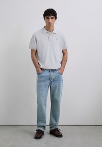 Polo de color gris claro con cuello, detalle del logo en azul, combinado con unos vaqueros de mezclilla azul claro y zapatos de cordones marrón oscuro.