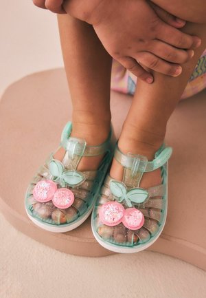 Malé dieťa má na nohách priehľadné svetlozelené sandále s ružovými ozdobami v tvare čerešní, sedí so založenými rukami na skrížených nohách.