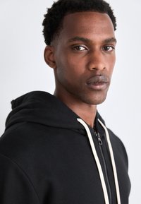 Sudadera negra con cremallera y capucha ajustable, con cordones de color crema y una textura suave, llevada por una persona que mira directamente a la cámara.