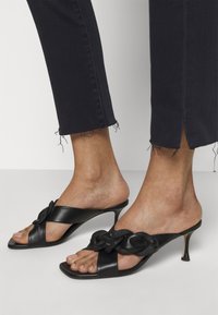 Pies con mules negras de tacón bajo con tiras trenzadas y dedos abiertos, combinados con pantalones negros recortados con dobladillo deshilachado.