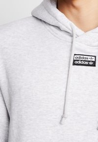 Ljusgrå Adidas-hoodie med en framsida där det finns en bröstficka med Adidas-logotypen och varumärket i svart och vitt.