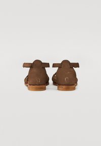 Sandales en suede marron avec une sangle à la cheville à l'arrière, un petit talon en bois et des découpes sur les côtés. Semelle plate pour le confort.