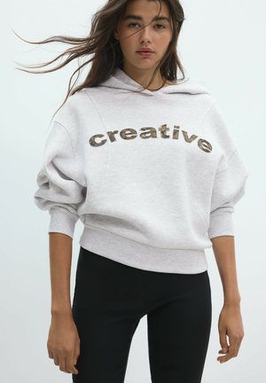 Jeune femme aux longs cheveux portant un sweat à capuche gris clair avec le texte "creative" et un pantalon noir sur un fond uni.