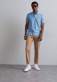 Light blue polo shirt, beige trousers, and white sneakers. Black sunglasses worn. Model stands against a neutral background.