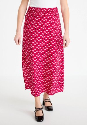 Frau trägt einen roten gemusterten Midirock und schwarze Mary Jane Absatzschuhe, steht vor einem schlichten weißen Hintergrund.