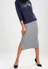 Mujer vestida con un sudadera azul marino con el colorido texto "ESPRIT", una falda midi gris con una abertura lateral y zapatos de tacón negros, de pie.