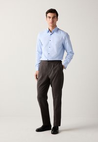 Chemise bleu clair à manches longues à boutons, pantalon à carreaux foncés et chaussures habillées noires. Le modèle se tient avec une main dans la poche sur un fond neutre.