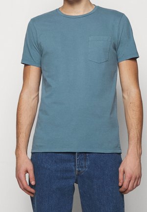 Mann trägt ein schlichtes, gedämpftes türkisfarbenes T-Shirt mit kurzen Ärmeln, Rundhalsausschnitt, einer kleinen linken Brusttasche und blaue Jeans aus Denim.
