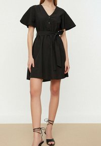 Robe noire évasée avec un décolleté en V, des manches courtes volantées, cinq boutons sur le devant et une ceinture en tissu ; associée à des talons noirs à lanières.