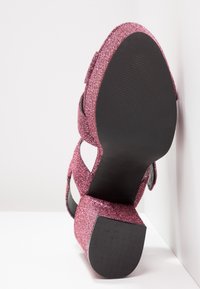 Sandale à talon bloc pailletée rose à bout ouvert, semelle texturée noire et lanières croisées, positionnée droite contre un mur blanc.
