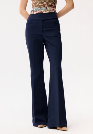 Kobieta ubrana w granatowe spodnie z wysokim stanem i szerokimi nogawkami, z przednimi kieszeniami, oraz beżowe, spiczaste buty, stoi na jednolitym tle.