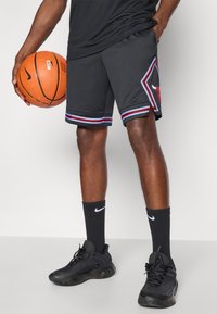 Juodos sportinės šortai su raudonais ir mėlynais akcentais, turintys logotipo užrašą. Su jais derinamos juodos "Nike" kojinės ir batai, laikoma krepšinio kamuolys.