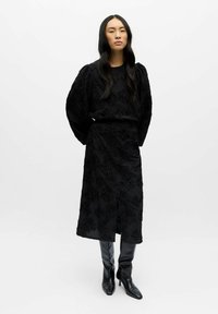 Kvinde med langt sort hår iført en sort tekstureret blomsterprint midi-kjole og sorte støvler med høje hæle, stående mod en ensfarvet baggrund.