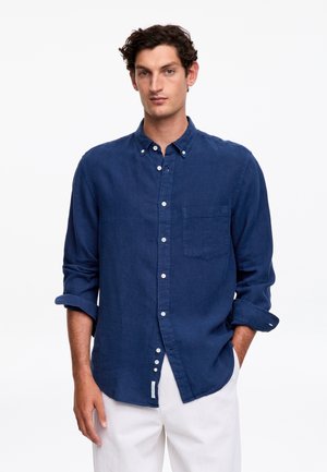 Mężczyzna stoi z rękami luźno opuszczonymi, ubrany w granatową koszulę na guziki z długim rękawem i białe spodnie na jednolitym białym tle.