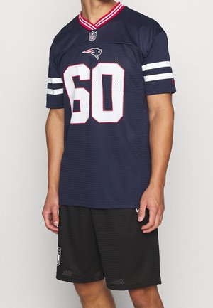 Mężczyzna ubrany w ciemnoniebieską koszulkę NFL New England Patriots z numerem 60 oraz czarne sportowe spodenki, stojący na jednolitym tle.